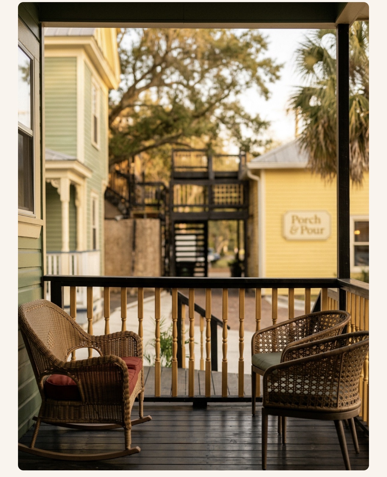 View from a shaded porch with wicker chairs, looking across to the sage-green Heron House and the Porch & Pour sign on the yellow Victorian