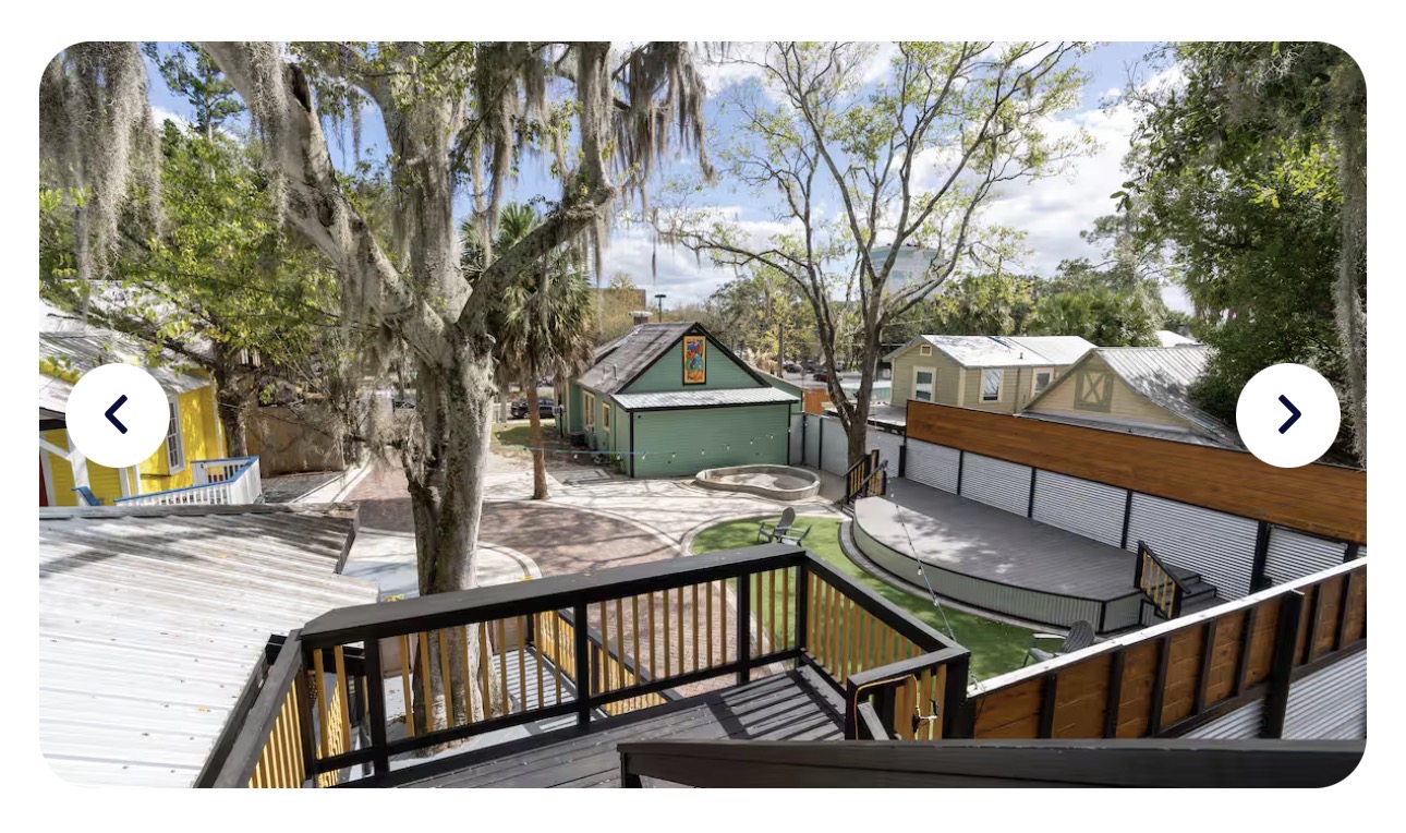Looking down from the Treehouse deck across the brick courtyard: live-oak canopy, yellow Porch & Pour at left, sage Heron House across, fire pit and Adirondack chairs