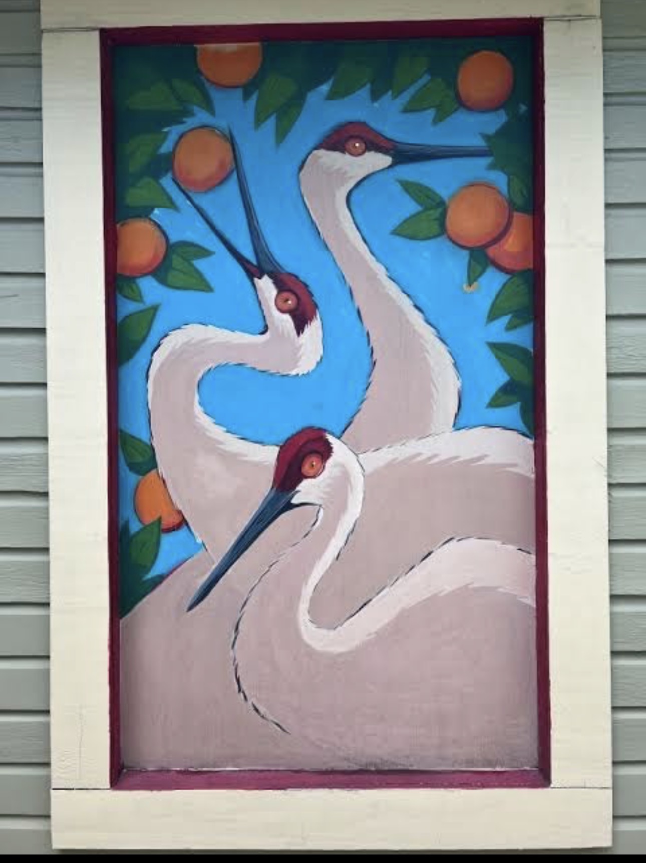 A hand-painted folk-art panel of three sandhill cranes among Florida orange branches, mounted on the sage Victorian's siding — a one-of-a-kind window the artist made for the village